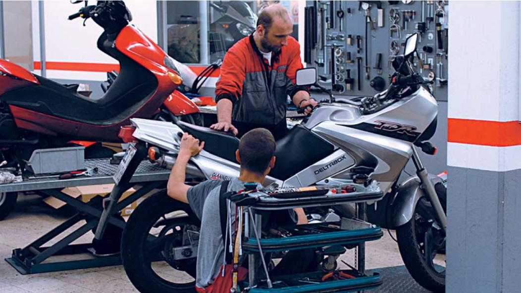 Bild einer Werkstatt, in der zwei Personen an einem Motorrad arbeiten.